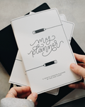 A person's hands holding a white monthly themed planner with black text reading 'my planner' and pencil illustrations, with other black planners stacked underneath on a gray concrete surface.