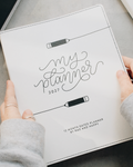 A person's hands holding a white 2027 monthly planner with black script lettering that reads 'my planner 2027' and pencil illustrations, set against a gray tabletop background.