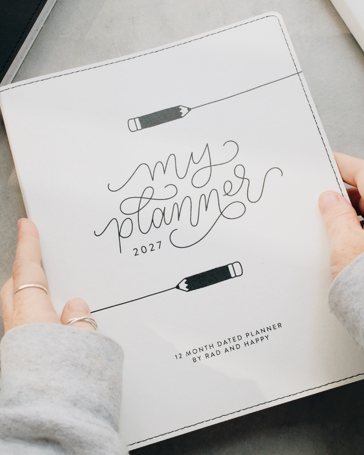 A person's hands holding a white 2027 monthly planner with black script lettering that reads 'my planner 2027' and pencil illustrations, set against a gray tabletop background.
