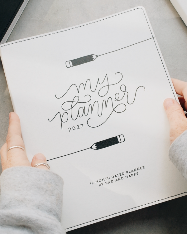 A person's hands holding a white 2027 monthly planner with black script lettering that reads 'my planner 2027' and pencil illustrations, set against a gray tabletop background.