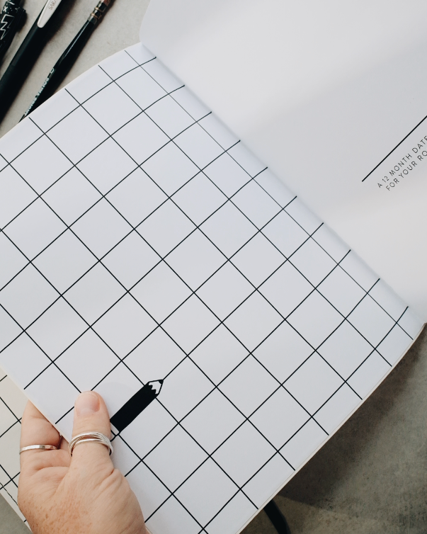 A hand with rings holds a white grid-bound notebook open on a gray concrete surface, with pens and a laptop visible in the background.
