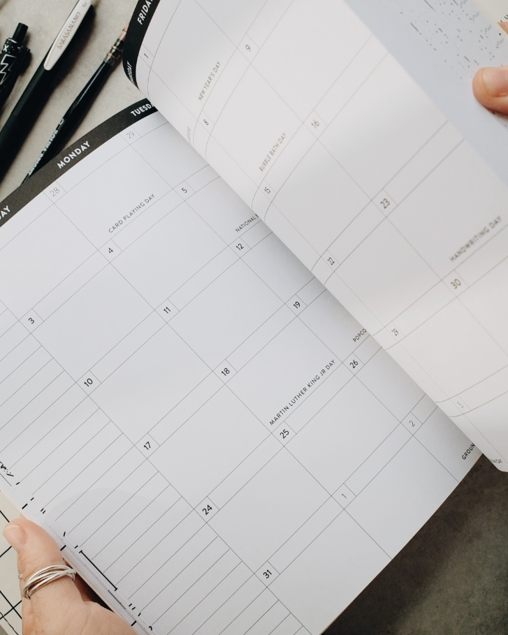 A person's hands are shown flipping through the pages of a black and white planner on a gray concrete surface, with pens scattered nearby.