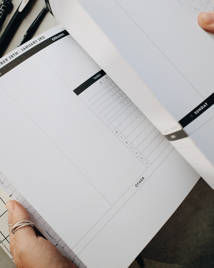 A close-up of a hand flipping through the pages of a leather-bound planner on a gray surface, with black pens nearby and handwritten notes visible on the page.