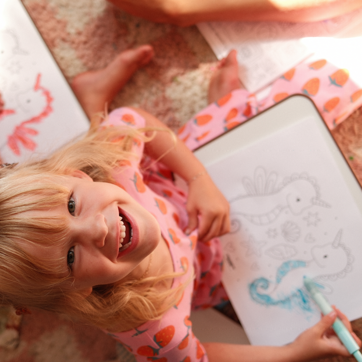 A young girl with blonde hair smiles while coloring in a white drawing pad with a blue crayon, lying on a carpeted floor. She is wearing a pink strawberry-patterned dress, and other drawing pads are visible nearby.