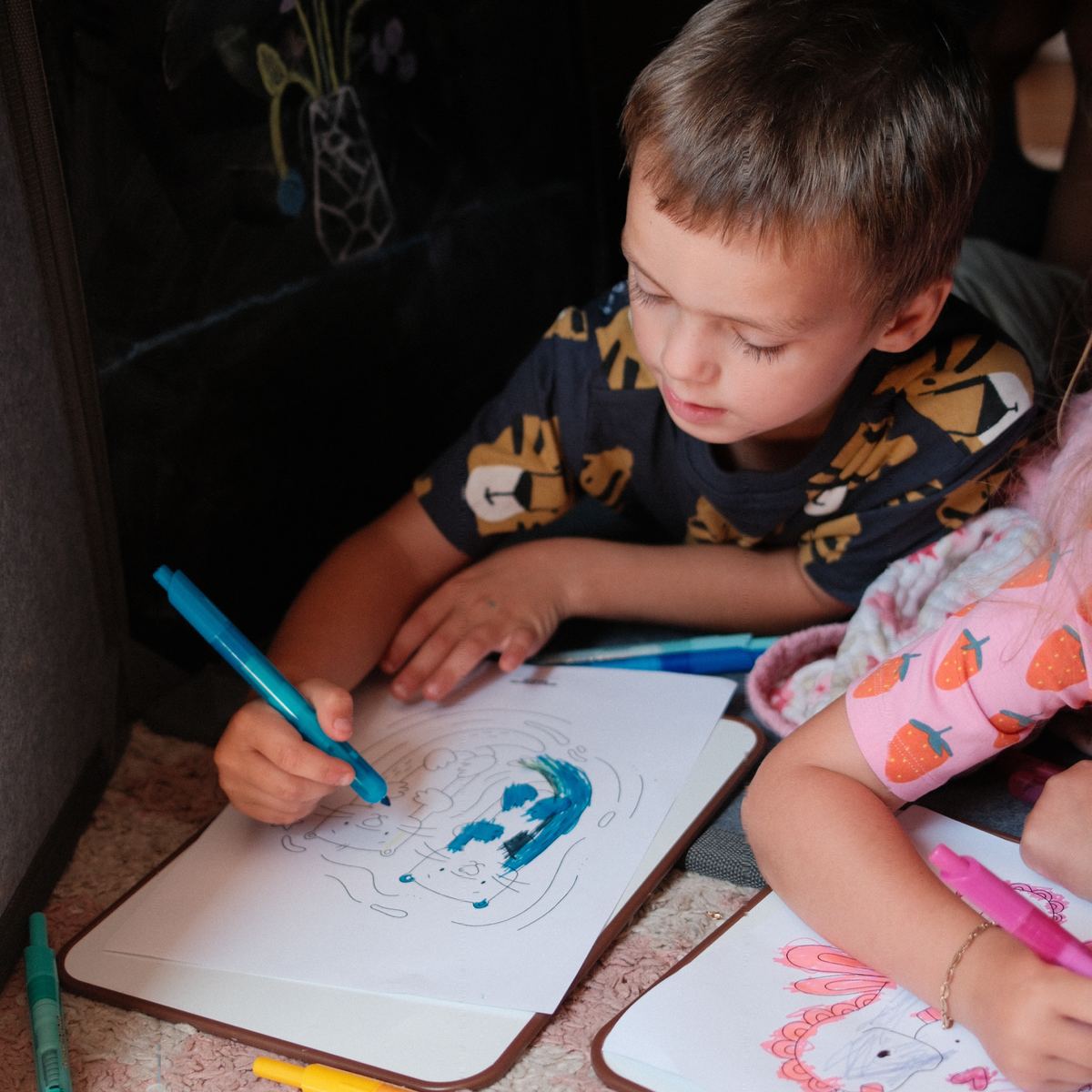 A young boy in a dark navy t-shirt with a lion graphic is drawing on a white sheet of paper with a turquoise marker. A young girl in a pink strawberry shirt is partially visible on the right, holding a pink marker.