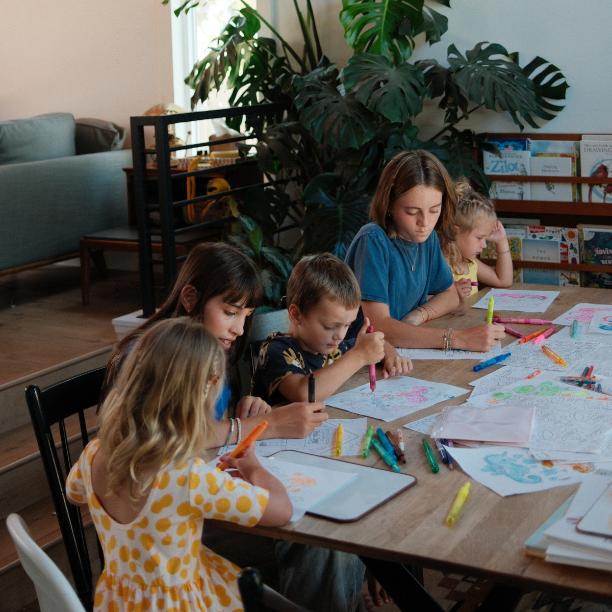 Four children and an adult woman sitting around a wooden table, drawing and coloring on paper with crayons and markers in a bright, plant-filled home office or library.