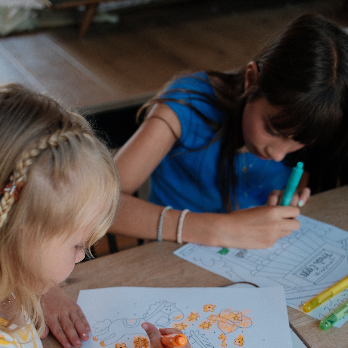 Two young girls coloring on white paper at a wooden table, one in the foreground with blonde braided hair holding an orange marker, and the other in a blue shirt writing with a turquoise marker.