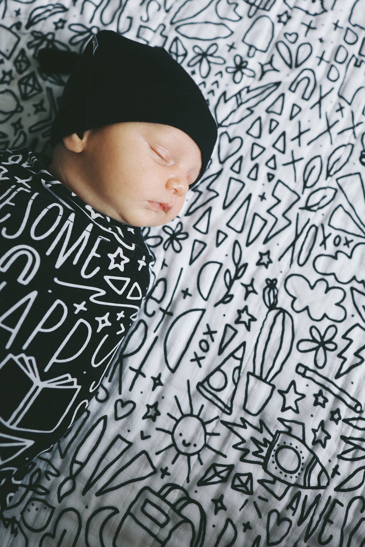 A baby in a beanie swaddled, laying on top of the quilt