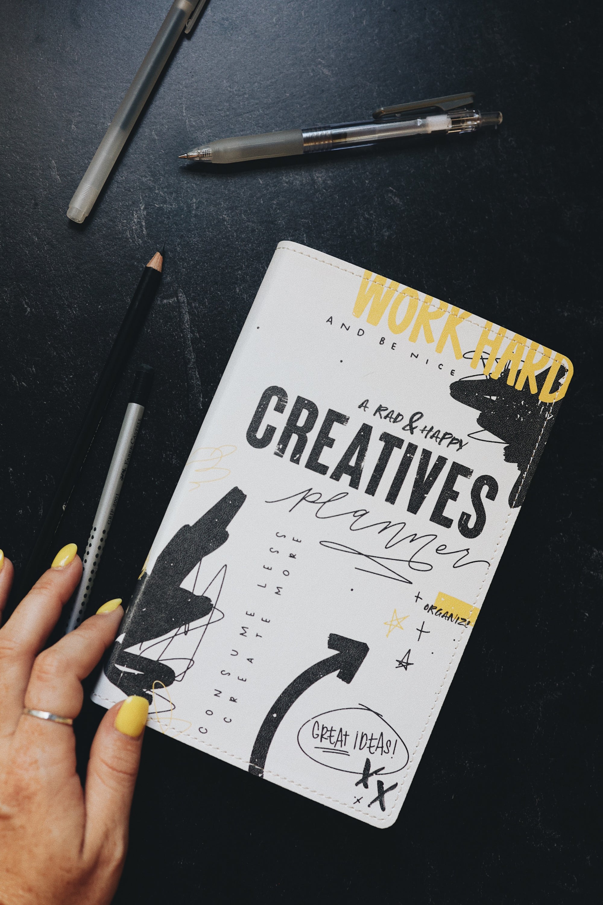 Artistic shot of a hand touching the creative planner on a desk with pens around it.