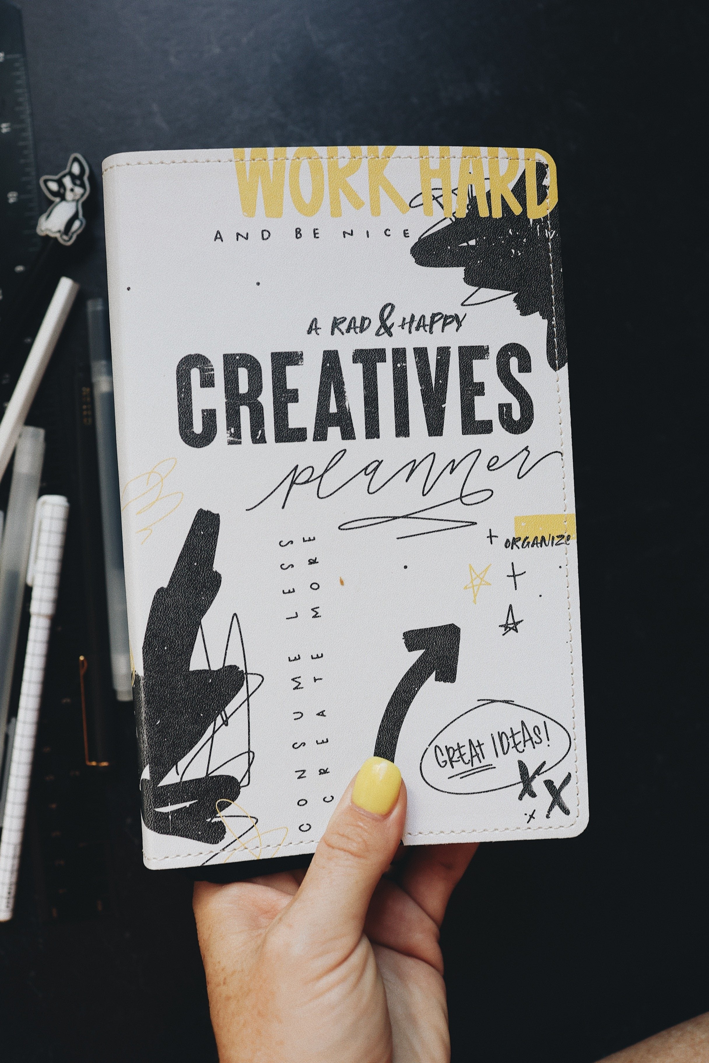 A hand holding the creative planner to show the cover. Desk top in background with pens and a ruler.