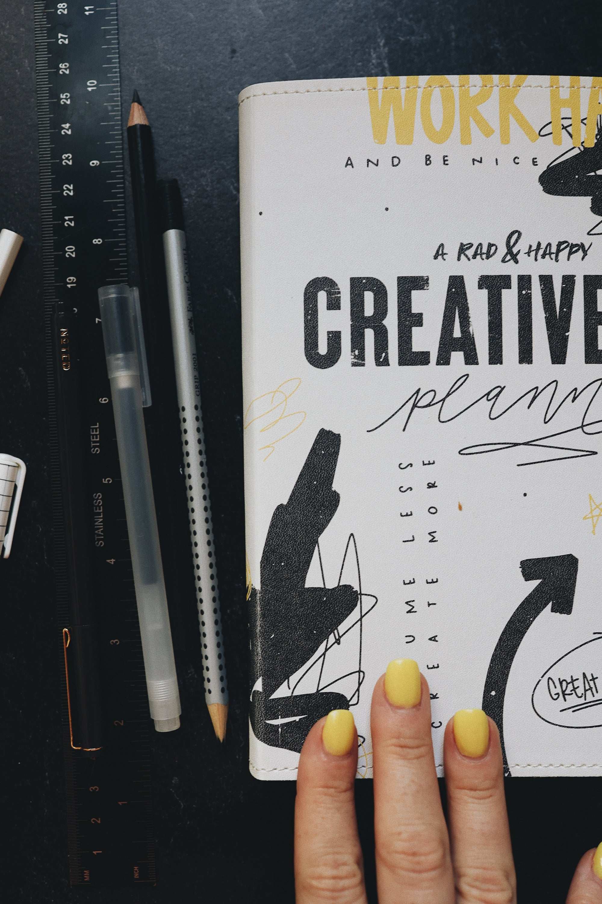 Artistic shot of the creative planner next to a ruler and pens, with three fingers on the bottom of the cover.