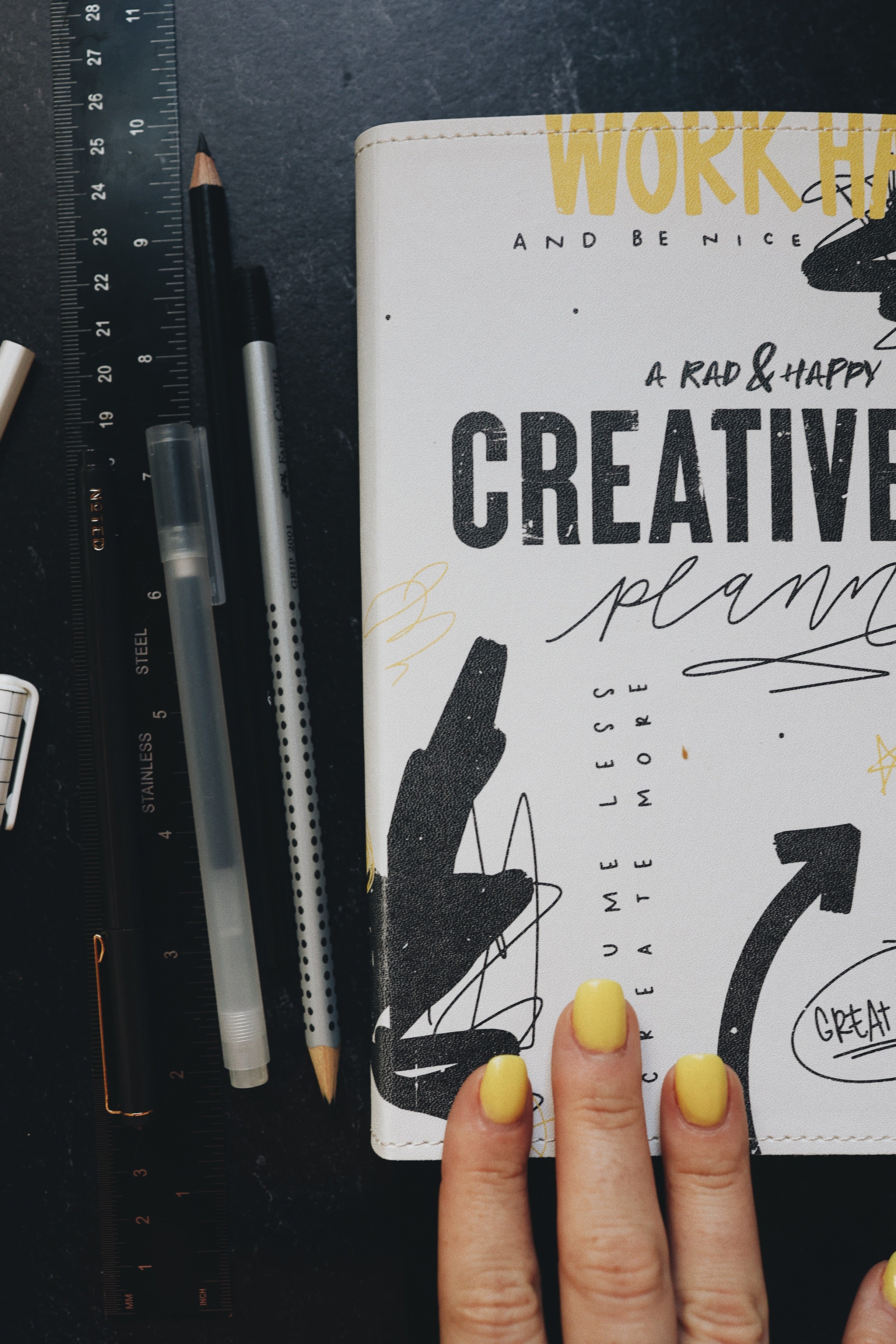 Artistic shot of the creative planner next to a ruler and pens, with three fingers on the bottom of the cover.